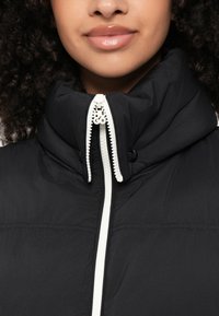 Schwarze Zip-Jacke mit hohem Kragen, ausgestattet mit einem weißen Reißverschluss und Logodetail, glatte Textur und keine sichtbaren Muster oder Akzente.