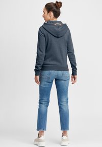 Mørk grå hettegenser med en avslappet passform, justerbar hette og brune detaljer. Kombinert med lyseblå jeans og hvite joggesko. Myk tekstur.