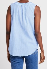 Blusa senza maniche azzurra e bianca a righe verticali con scollatura arricciata, indossata con jeans in denim blu, vista da dietro.