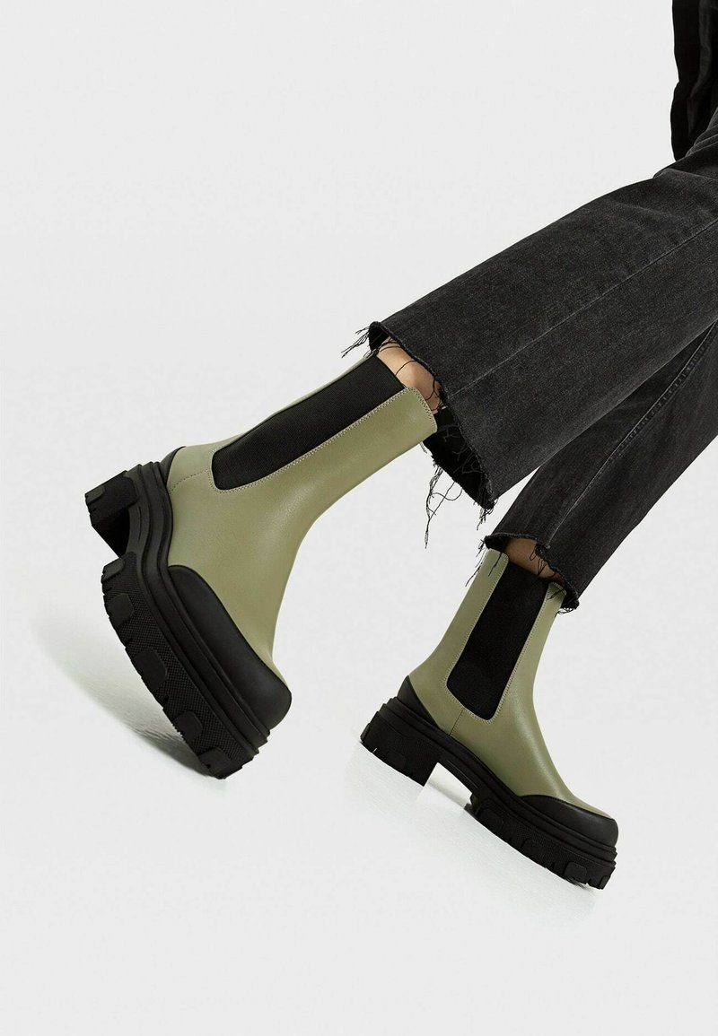 Bottines kaki à semelles épaisses noires, portées avec un jean noir à ourlet effiloché, sur un fond clair uni.