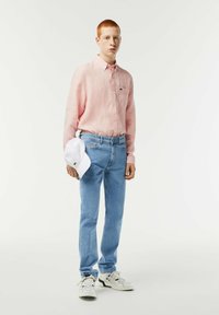 Camicia rosa chiaro a maniche lunghe, jeans in denim blu, cappellino bianco tenuto in mano e sneakers bianche con dettagli neri. Design semplice e informale.