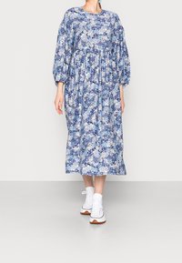 Žena v volné modré květinové midi šaty s dlouhými nabíranými rukávy a bílými vysokými teniskami, stojící na jednoduchém světlém pozadí.