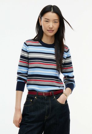 Ung kvinde med langt mørkt hår iført en stribet sweater med blå, grå, røde og sorte striber, mørke jeans og et rødt bælte, stående med hånden i lommen.