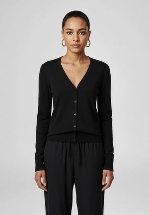 Žena s krátkými vlasy nosí černý zapínaný kardigan s výstřihem do V a volné černé kalhoty, s pozlacenými kruhovými náušnicemi, stojí před jednoduchým pozadím.