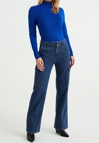 Blå långärmad tröja med hög nacke och knappar på ena axeln, kombinerad med höga denim cargo-byxor med sidofickor.