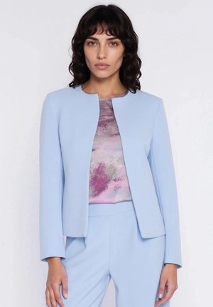 Kobieta ubrana w jasnoniebieską, dopasowaną marynarkę i spodnie oraz bluzkę z abstrakcyjnym wzorem w odcieniach różu i fioletu, stojąca na tle jednolitego białego tła.