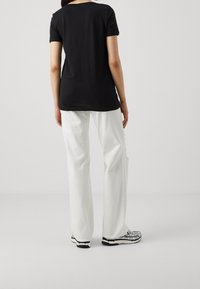 Person som bär en svart kortärmad topp, lösa vita byxor med en sidoklyfta och vita sneakers med svart och grå detaljer, stående med ryggen vänd.