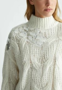 Maglione bianco lavorato a maglia con colletto alto a coste, caratterizzato da un design a cavo testurizzato e decorazioni floreali in paillettes sulle spalle.
