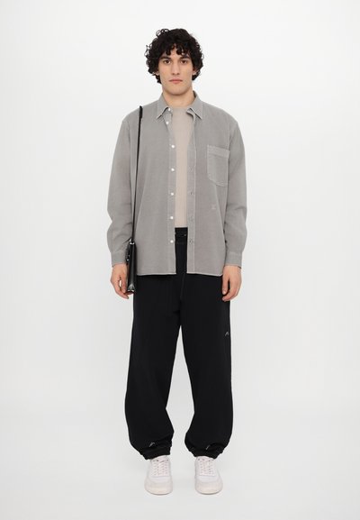 Chemise grise à boutons avec poche, haut beige en dessous, pantalon noir ample avec poignets en élastique, et baskets blanches. Sac bandoulière noir.