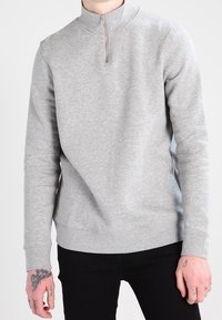 Grå zip hoodie med hög krage, mjukt material, långa ärmar och ribbad fåll. Har synliga tatueringar på händerna.