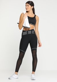 Svart sport-bh og høyt liv leggings med gjennomsiktige paneler. Leggingsene har teksten "JUST DO IT". Modellen holder et hvitt håndkle og har på seg grå sneakers.