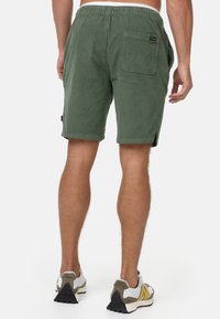 Grüne Cordshorts mit einem elastischen Bund, einer Gesäßtasche und seitlichen Schlitzen am Saum, von hinten gezeigt.