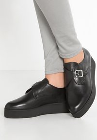 Chaussures noires en cuir avec un bout rond, détail de boucle et une épaisse semelle noire compensée, associées à un pantalon ajusté gris clair.