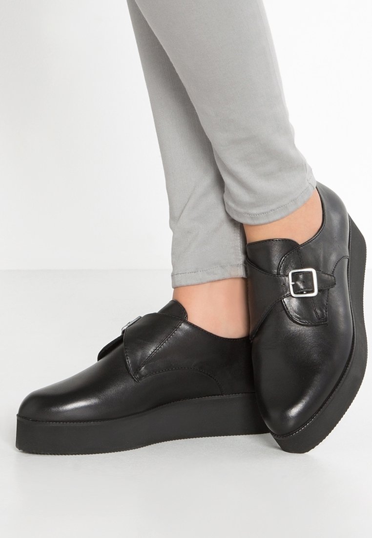 Chaussures noires en cuir avec un bout rond, détail de boucle et une épaisse semelle noire compensée, associées à un pantalon ajusté gris clair.