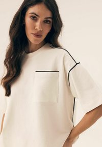 Biały, luźny T-shirt wykonany z miękkiego materiału, z krótkimi rękawami, kieszenią na piersi oraz kontrastowym czarnym wykończeniem na szwach.