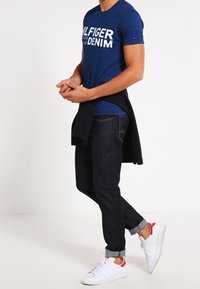 Námořnická modrá tričko s bílým nápisem "TOMMY HILFIGER DENIM", tmavé džíny a bílé tenisky s červenými akcenty; ukázka neformálního outfitu.