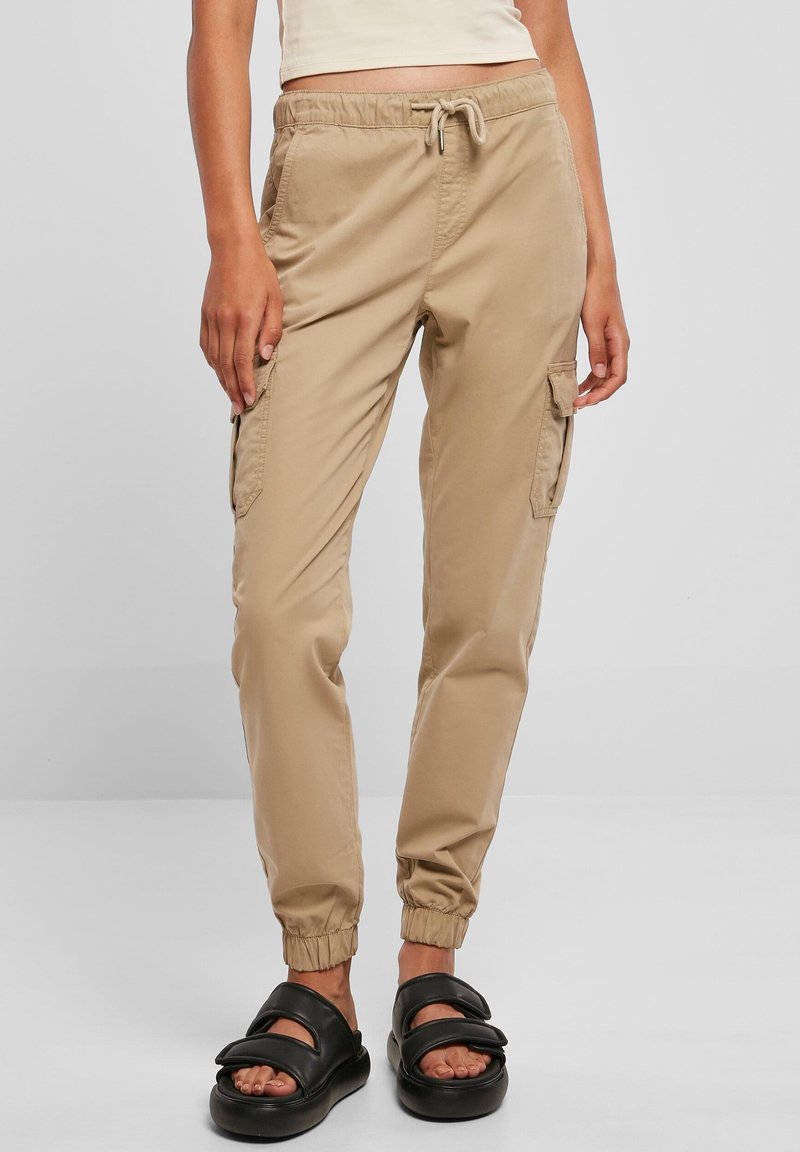 Beżowe spodnie cargo z elastycznymi mankietami, regulowanym paskiem ze sznurkiem w talii oraz bocznymi kieszeniami. Noszone z czarnymi sandałami na ślizg.
