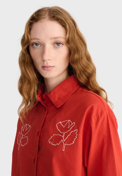 Jeune femme aux longs cheveux roux ondulés porte une chemise rouge à boutons ornée d'une broderie blanche de feuilles sur la poitrine sur un fond uni.