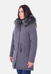 Cappotto imbottito grigio con colletto in pelliccia sintetica, chiusura con zip, vita regolabile con cordino e due tasche laterali. La superficie è liscia con una finitura morbida.