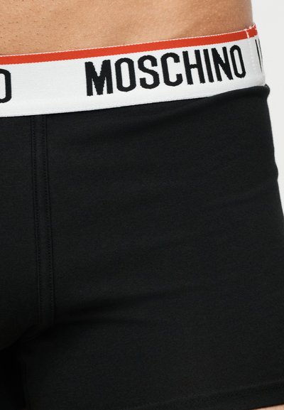 Μαύρα μποξεράκια με λευκή μέση που έχει μια κόκκινη ρίγα και το όνομα της μάρκας "MOSCHINO" με έντονα μαύρα γράμματα.
