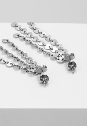 Boucles d’oreilles pendantes en chaîne métallique argentée avec maillons circulaires et attaches à tige sur une surface blanche.