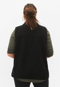 Gilet en polaire noir avec un col haut, texture douce et coupe décontractée. Superposé sur un haut à manches longues olive. Design simple.