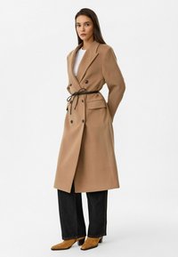 Dubbelzijdige camelkleurige jas met een strikou plooi, ontworpen met een kraag, zijzakken en gecombineerd met donkere denim en beige enkellaarsjes.