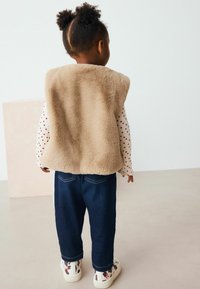 Beige fuskpälsväst över en vit långärmad tröja med röda prickar, kombinerat med mörkblå jeans och mönstrade sneakers, sedd bakifrån.