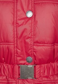 Chaqueta acolchada roja con botones a presión plateados y una hebilla de cinturón metálica con un acabado texturizado. Patrón acolchado en toda la tela.