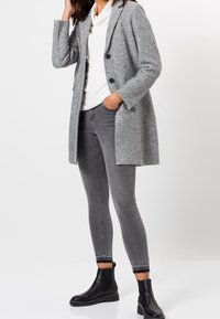 Szary płaszcz z mieszanki wełny o dopasowanym kroju, z dwoma guzikami i bocznymi kieszeniami, noszony na białym golfie i zestawiony z szarymi dżinsami oraz czarnymi botkami do kostki.