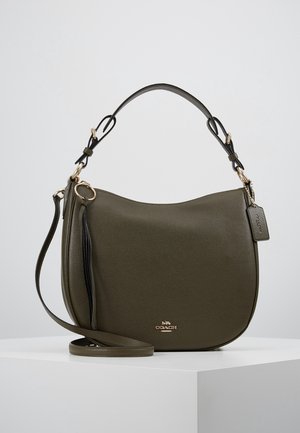 Oliwkowa skórzana torebka Coach z regulowanym paskiem na ramię, złotymi okuciami i wiszącą skórzaną zawieszką, ustawiona na białym piedestale ekspozycyjnym.
