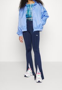 Ljusblå satinjacka med elastiska ärmar, mörkblå utsvängda leggings med sidussprund, och vita sneakers med färgglada detaljer.