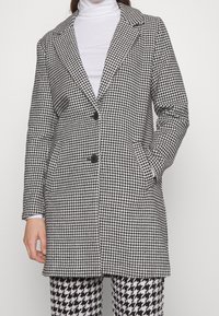 Abrigo largo de patrón de espiga en blanco y negro, con cuello con muesca, dos botones y bolsillos laterales. Tejido suave con un corte entallado.