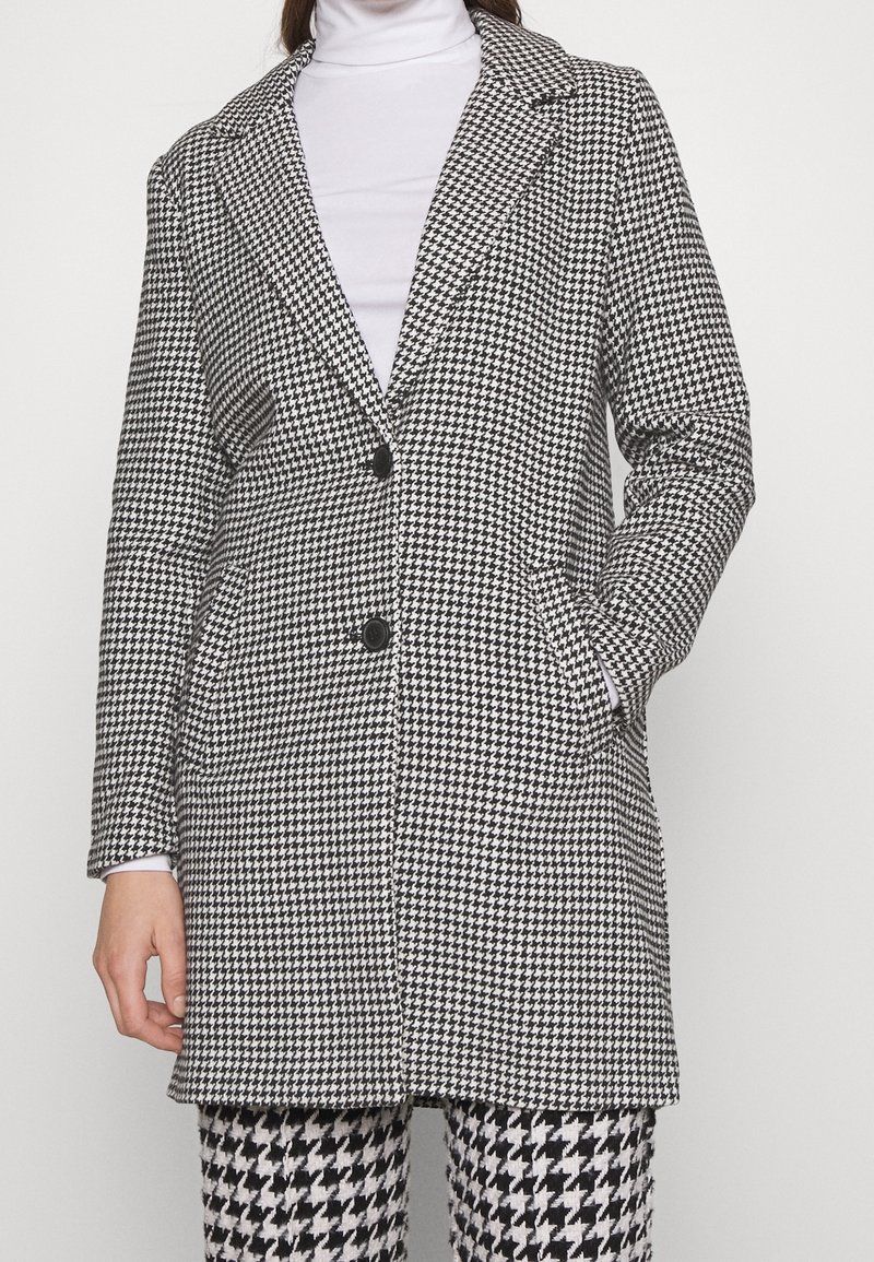 Abrigo largo de patrón de espiga en blanco y negro, con cuello con muesca, dos botones y bolsillos laterales. Tejido suave con un corte entallado.