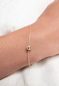 Złoty bransoletka z delikatnym łańcuszkiem i centralną kulkową perełką. Powierzchnia jest gładka i lśniąca w kontakcie ze skórą.