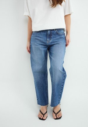 Femme portant un jean bleu droit avec des ourlets bruts, un haut blanc oversize à manches courtes et des sandales noires à bout carré.