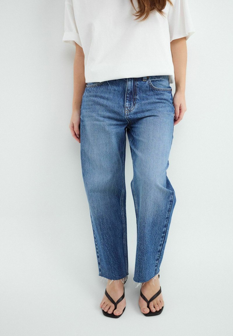 Femme portant un jean bleu droit avec des ourlets bruts, un haut blanc oversize à manches courtes et des sandales noires à bout carré.