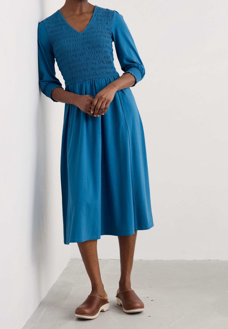 Femme portant une robe bleue mi-longue avec corsage froncé et manches trois quarts, debout contre un mur blanc, sabots marron aux pieds.