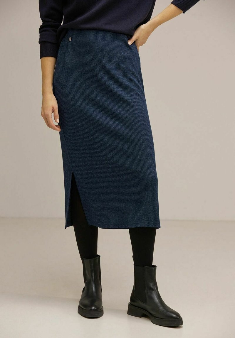 Femme portant une jupe midi côtelée bleu foncé avec une fente latérale, des collants noirs, des bottines en cuir noir et un pull bleu marine.