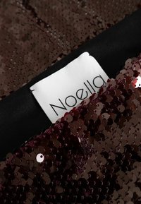 Burgundi színű flitteres anyag fényes textúrával, fekete béléssel és egy látható fehér címkével, amelyen fekete betűkkel az "Noella" felirat szerepel.