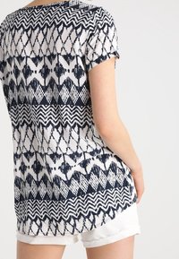 Kortärmad toppen i vitt med marinblå geometriska mönster, med rund halsringning och en längre bakre fåll. Tyget verkar vara lätt.