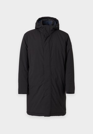 Homme portant une casquette marron, un manteau à capuche noir, un pull crème avec un logo circulaire, un pantalon beige à carreaux, des bottes noires, debout avec une main dans la poche.