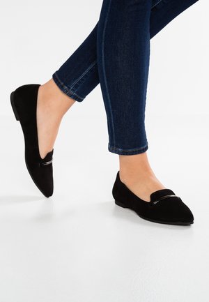 Person iført mørkeblå tætsiddende jeans og sorte loafers i ruskind med en lille metaldetalje på en hvid gulv.