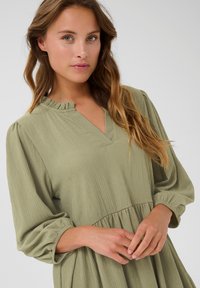 Grünes, strukturiertes Kleid mit einem rüschierten V-Ausschnitt, langen, gepufften Ärmeln und einem gerafften Bund. Der Stoff hat eine weiche, subtile Rippoptik.
