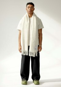 Écharpe blanche en tricot avec des franges, portée sur une chemise claire et un pantalon plissé noir. Des baskets vertes complètent la tenue.