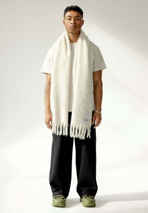 Écharpe blanche en tricot avec des franges, portée sur une chemise claire et un pantalon plissé noir. Des baskets vertes complètent la tenue.
