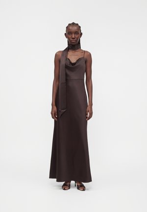 Femme aux cheveux tressés porte une robe nuisette longue marron foncé à fines bretelles et une écharpe assortie nouée autour du cou.
