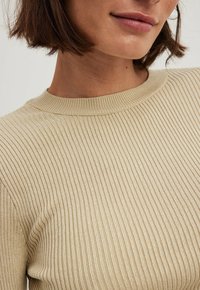 Pull en maille côtelée beige avec un col rond, une coupe ajustée et une texture lisse avec des lignes verticales.