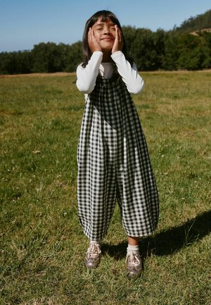 Young girl wearing black-and-white checkered overalls and white long sleeve shirt stands on grass with eyes closed, hands gently on cheeks.