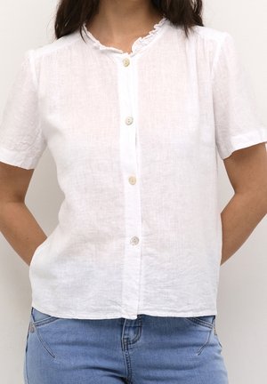 Blouse blanche à manches courtes en lin léger, avec un col ruffled et cinq boutons sur le devant, associée à un jean bleu.